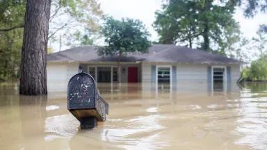 Floodwaters rise near the windows of a single-story house, with a mailbox almost underwater in the foreground.
