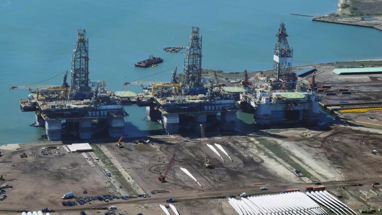 Three large oil rigs sit in water just next to a mostly paved port area.