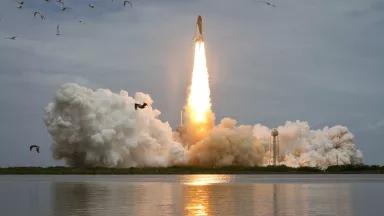 A space shuttle launches from the ground, burning fuel and creating plumes of smoke.