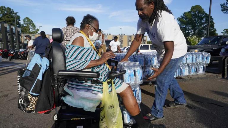In a city parking lot, a man helps a woman in a wheelchair with a case of bottled water.