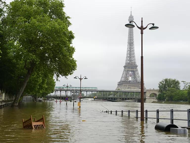 An outdoor chair floats upside in floodwaters with trees and the Eiffel Tower in the background