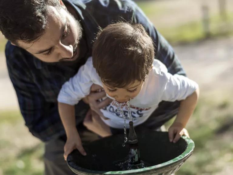 A father helping his young son drink from a water fountain.