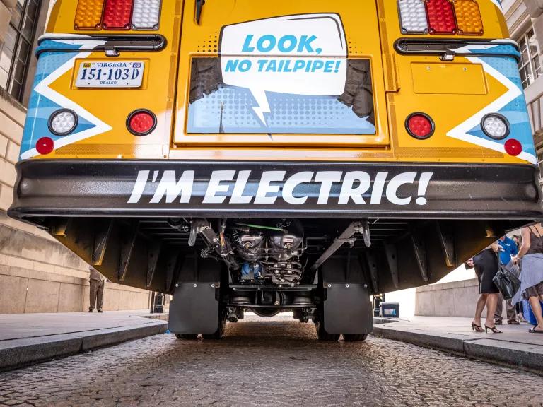 The back of yellow bus with the words "Look, no tailpipe! I'm electric!" painted on it