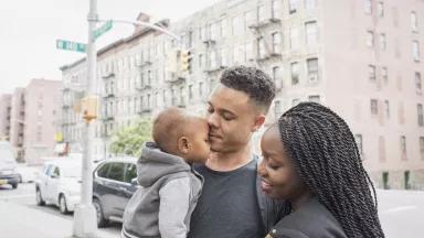 A Black woman stands next to a Black man who is holding a toddler on a city street with an apartment building in the background