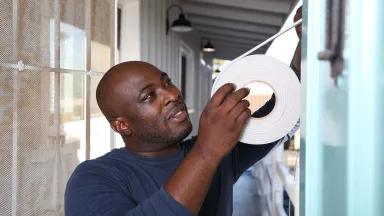 A Black man is applying a role of weather tape to an exterior door of a home. 