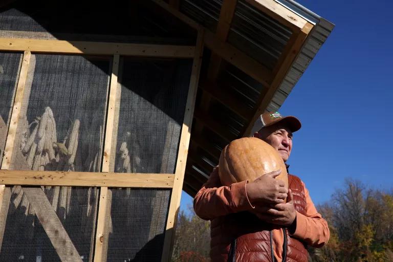 A man in a cap and outdoor vest in front of a wooden building holds a large squash