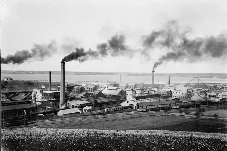 A black and white image of an industrial plant on the banks of a body of water, with black smoke rising from three smokestacks
