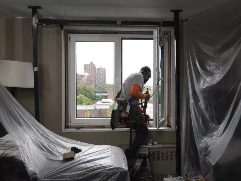 A worker stands at a window in a room with plastic sheeting hanging from the ceiling and covering furniture
