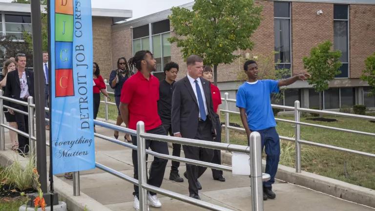 U.S. Secretary of Labor Marty Walsh, wearing a black suit and blue tie, walking in front of the Detroit Job Corps Center building with a person in a blue T-shirt on this right, pointing his finger at something in the distance, a person in a red collar T-shirt on his left, and a group of other people walking behind them