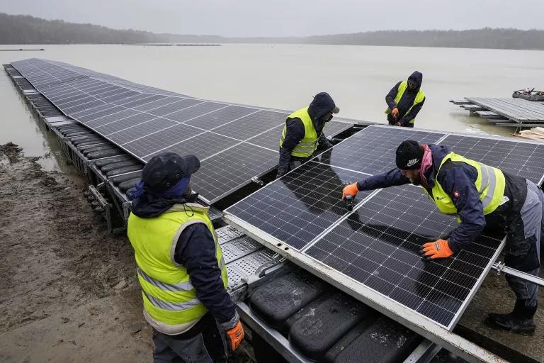 Workers move a large solar panel into place in a row on the shore of a lake 