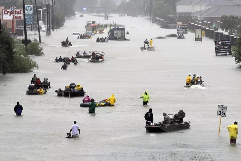 Multiple rafts and boats travel through floodwaters on a multi-lane roadway, along with people walking in the waist-high water