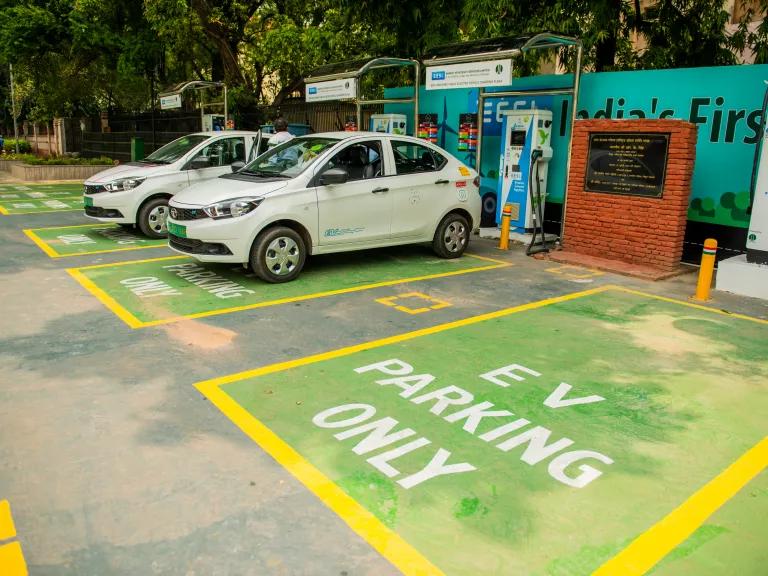A parking lot with green parking spots that have signs that read "EV parking only," with drivers charging their electric cars in New Delhi, India