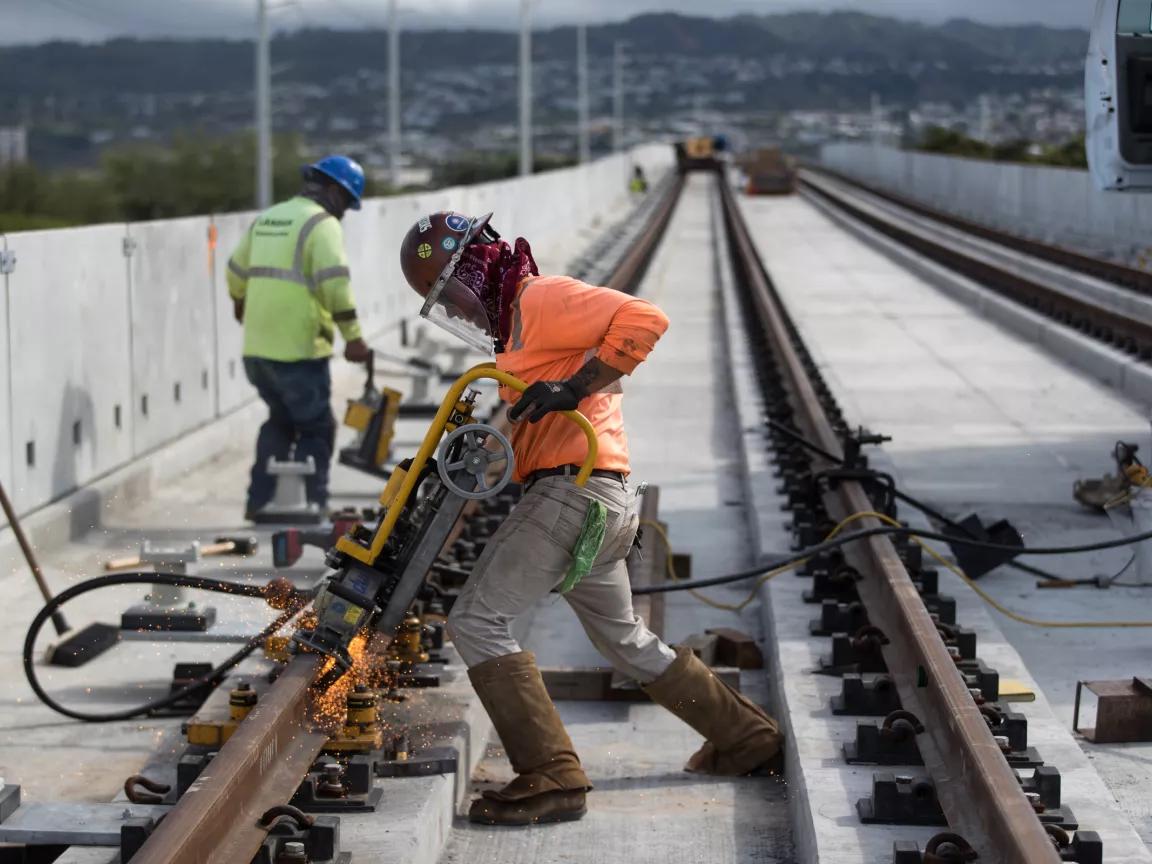 Two construction workers, with one in the forefront wearing an orange shirt, at a construction site for the Honolulu Rail Project in 
