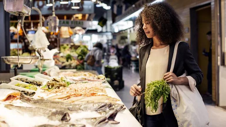 A Black woman holding a cloth shopping bag filled with produce is looking at fish on ice at a market.