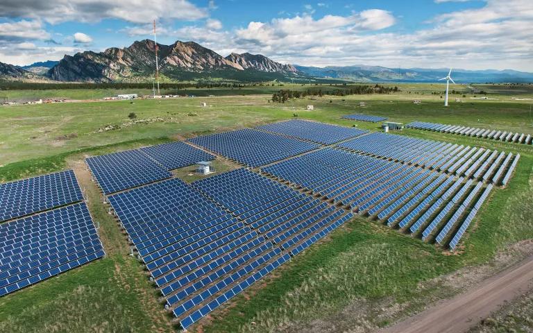 An array of solar panels in a large grassy field, with a wind turbine and mountains in the background