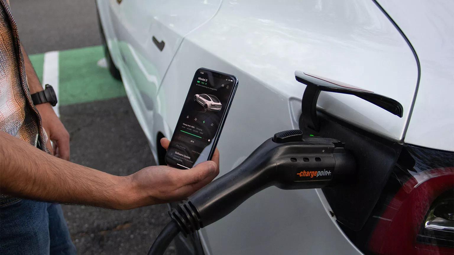 A person stands next to their Tesla and views its charging progress in a smartphone app