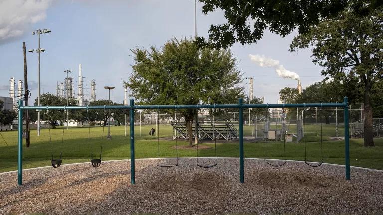 Plumes of smoke from an oil refinery rise behind a swing set in a children’s playground