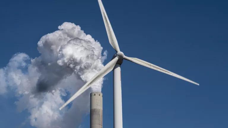 A wind turbine stands in front of a smokestack